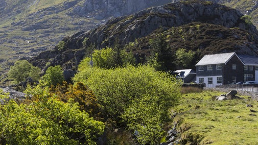 A view of Ogwen Cottage, Carneddau and Glyderau, Gwynedd, with Cwm Idwal Valley behind. The cottage is a large black and white building and sits beside a stream in a rugged valley.